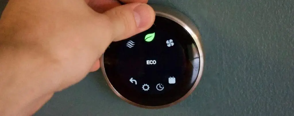 Hand turning a circular smart thermostat set to eco mode on a wall.