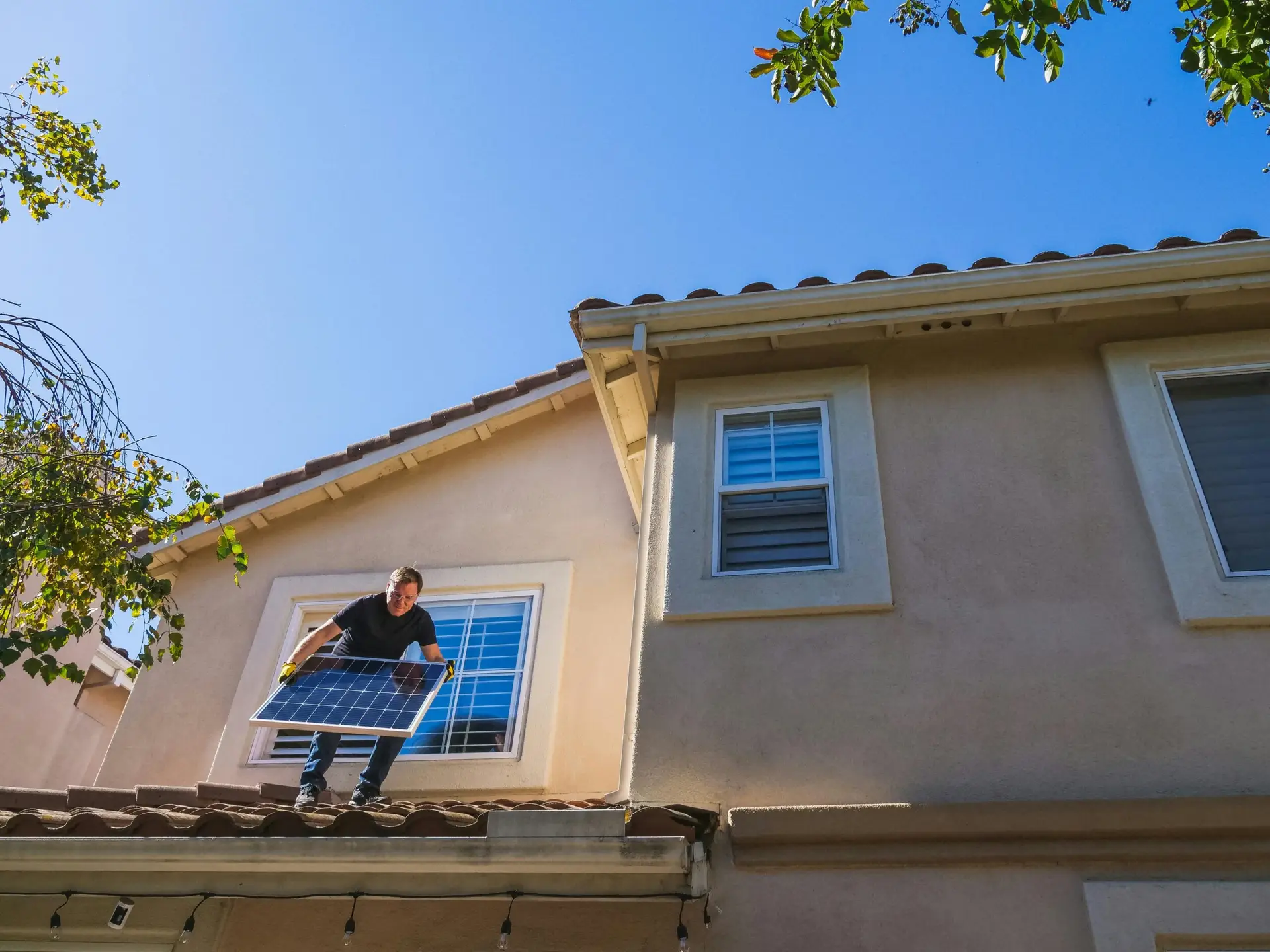 A man installing a solar panel on the tiled roof of a beige two-story house under a clear blue sky.