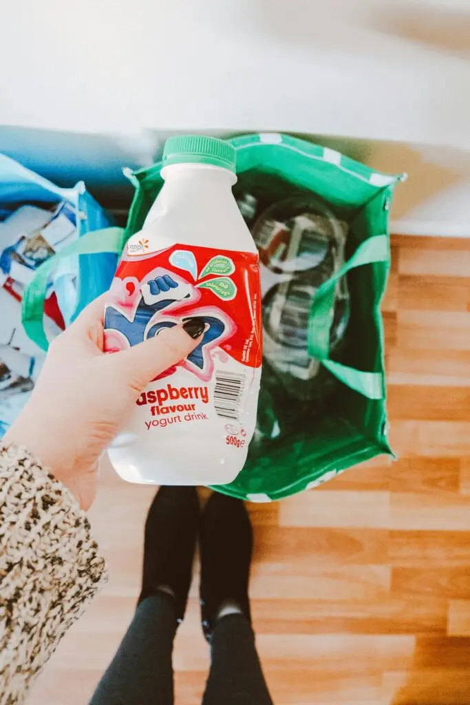 Person holding an empty yogurt drink bottle above a green recycling bag filled with other recyclables on a wooden floor.